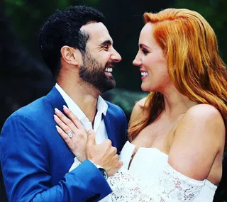 A couple smiling at each other, the woman in a white dress showing an engagement ring, outdoors.