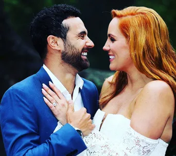 A couple smiling at each other, the woman in a white dress showing an engagement ring, outdoors.