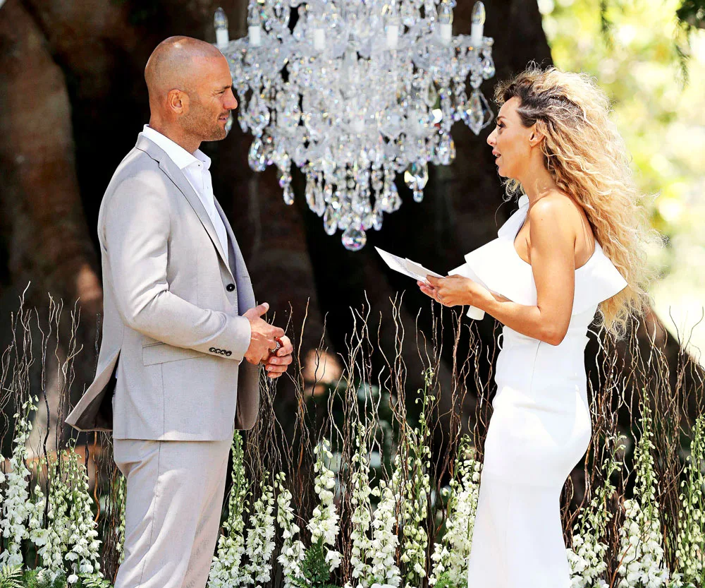Man in a gray suit and woman in a white dress exchanging vows outdoors under a chandelier.