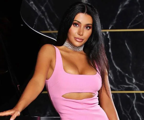 A woman in a pink dress with cutouts poses confidently, set against a dark marble background and a grand piano.