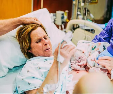 A woman in a hospital bed holds a newborn baby wrapped in a blanket, with a hand gently resting on her head.