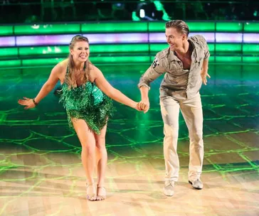 Dancers performing energetically on a lit stage, one in a green dress and the other in a beige outfit.