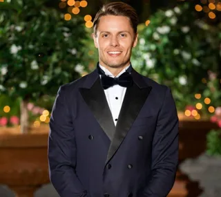 Man in formal tuxedo smiling in front of decorative garden lights.