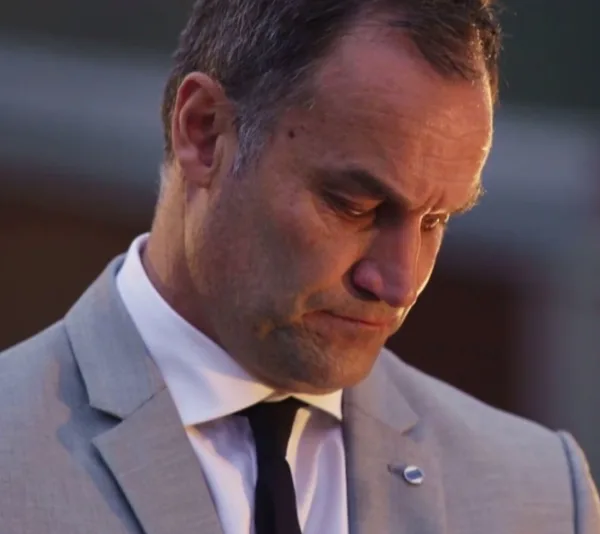A serious man in a gray suit and black tie looking down, with a blurred background.