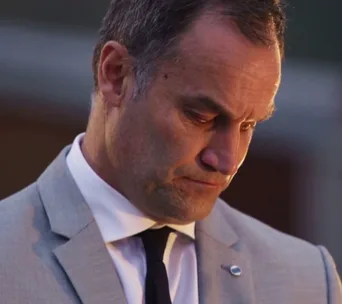 A serious man in a gray suit and black tie looking down, with a blurred background.