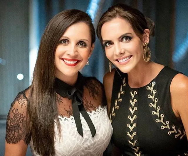 Two women smiling, dressed elegantly. One in a lace dress, the other in a black dress with gold accents, indoors.