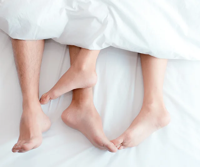 Feet of a couple intertwined under white bed covers, suggesting intimacy and relaxation.