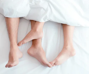 Feet of a couple intertwined under white bed covers, suggesting intimacy and relaxation.