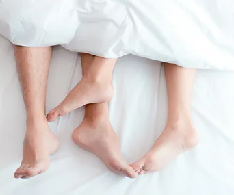 Feet of a couple intertwined under white bed covers, suggesting intimacy and relaxation.