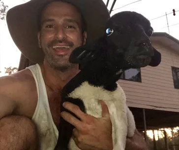 Man in a wide-brimmed hat holding a black and white lamb against a sunset backdrop with a house behind.