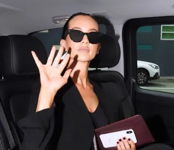 Individual in sunglasses waves from car, holding phone and maroon clutch.