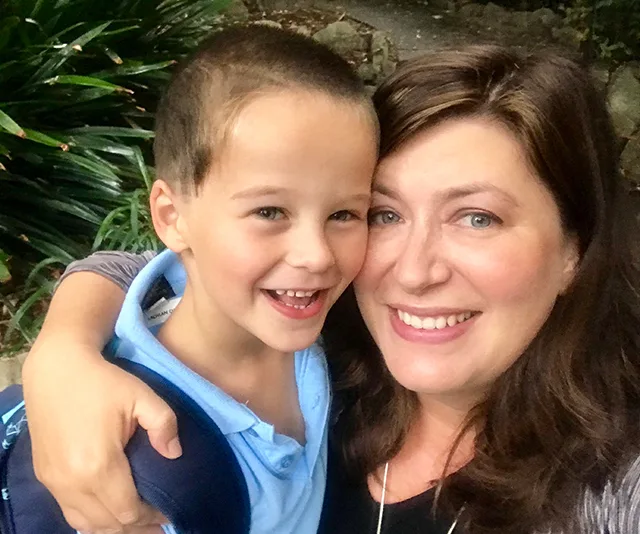 A woman and young boy smiling warmly, embracing outdoors with greenery in the background.