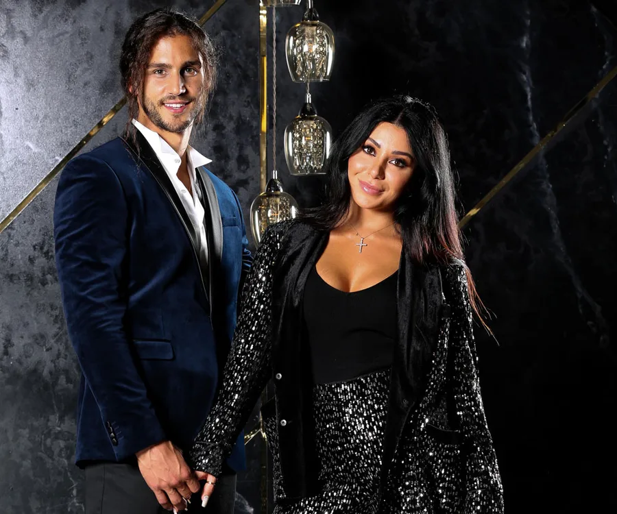 A couple poses elegantly in formal attire, holding hands with a backdrop of decorative pendant lights.