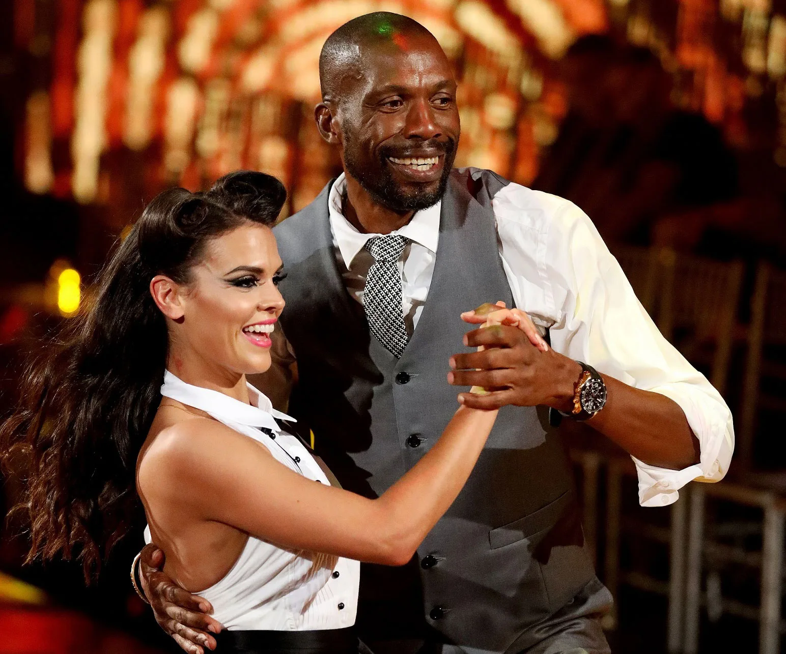 "Dancing couple smiling and holding hands on stage under warm lights."