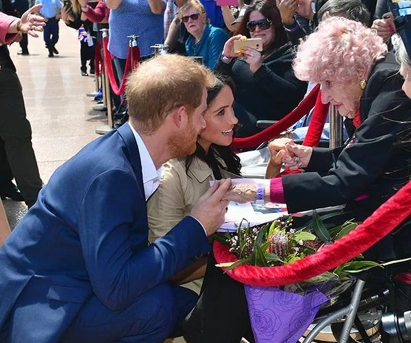 Two people kneeling, engaging with an elderly woman in a wheelchair, surrounded by onlookers and flowers nearby.