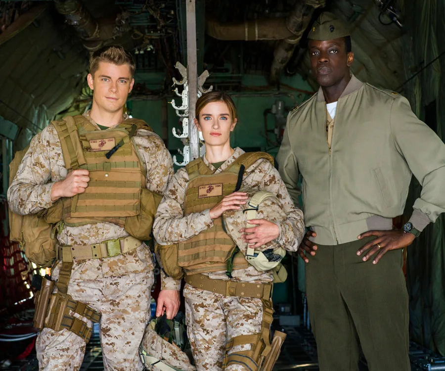 Three people in military uniforms standing inside an aircraft, two in camo gear with helmets, one in green jacket and cap.