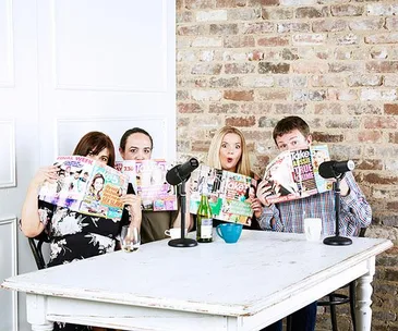 Four people at a table holding up magazines, with microphones and drinks in a cozy, brick-walled room.