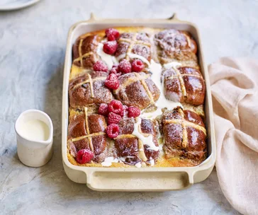 Baked hot cross buns with cream and raspberries in a dish, next to a small jug of cream.