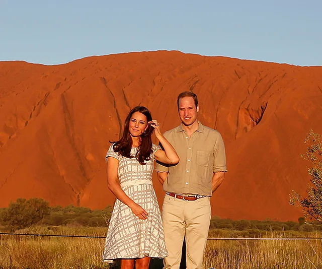 A couple stands in front of Uluru, a large red sandstone formation under a clear blue sky.