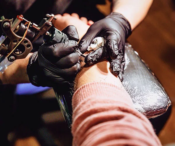 Tattoo artist with black gloves using a tattoo machine on a person's arm in a studio.