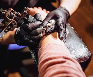 Tattoo artist with black gloves using a tattoo machine on a person's arm in a studio.