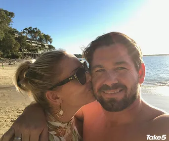 A couple taking a selfie on a sunny beach; the woman kisses the man while he smiles at the camera.