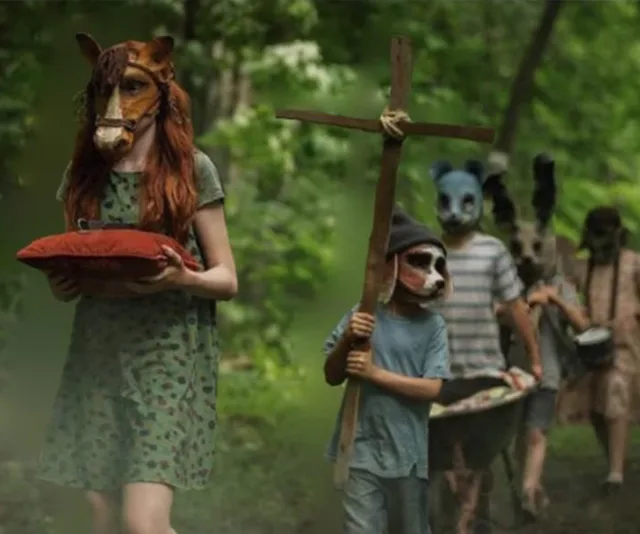 Children wearing animal masks walk through the woods, one with a cross, in a scene from "Pet Sematary" film.