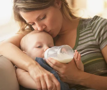 Mother cradles and feeds baby with a bottle while kissing baby's head.