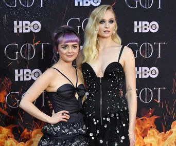 Two actresses pose on the Game of Thrones premiere red carpet, wearing black dresses with decorative elements.