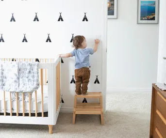 Toddler standing on a stool reaching for a light switch in a nursery with teepee wall decals and a crib nearby.