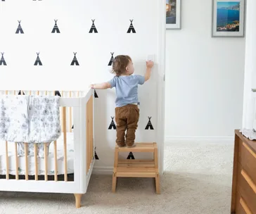 Toddler standing on a stool reaching for a light switch in a nursery with teepee wall decals and a crib nearby.