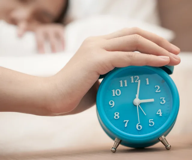 A hand reaching to turn off a blue alarm clock showing 7 o'clock, with a person sleeping in the background.