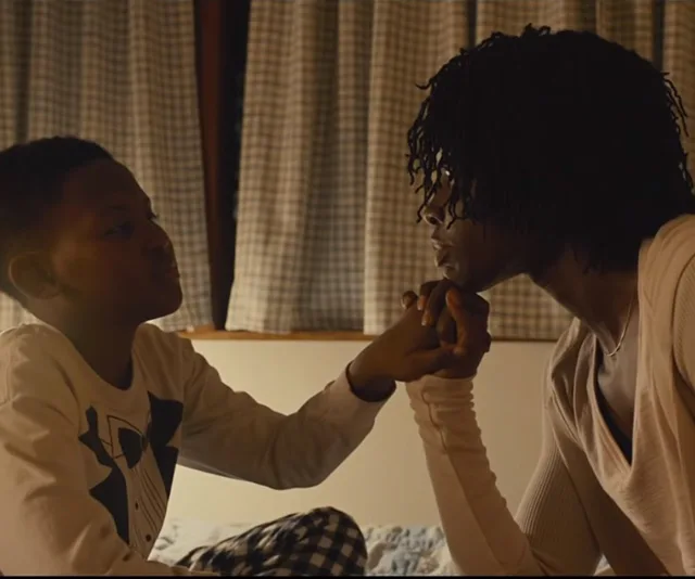 A boy and a woman sit facing each other, holding hands in a warm, intimate moment, indoors with checkered curtains in the background.