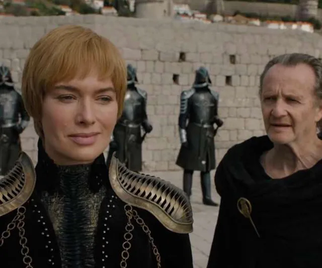 A blonde woman in armor stands with an older man, with guards in the background on a stone courtyard.