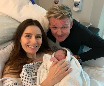 "Man and woman smiling at camera while woman holds newborn wrapped in a blanket at a hospital."