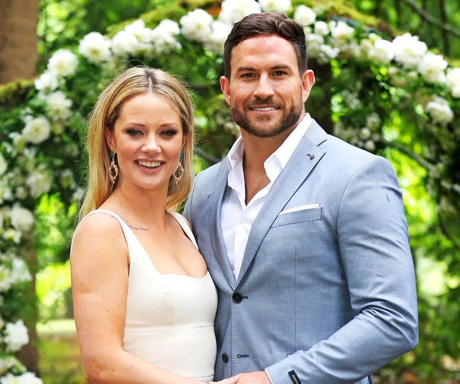 A couple smiling outdoors, woman in white dress, man in light blue suit, with floral greenery in the background.