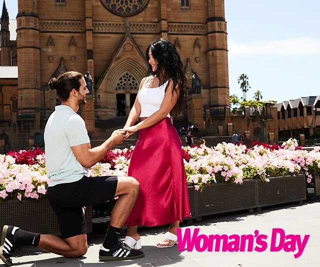 Man kneels holding woman's hands in front of a cathedral, surrounded by flowers, with "Woman's Day" logo.
