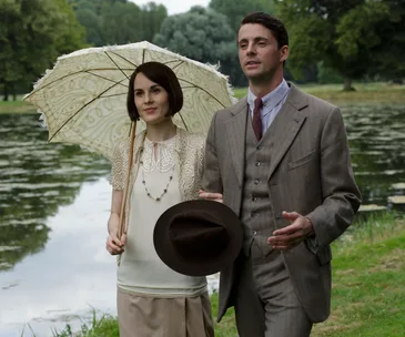 A man and woman in vintage attire walking by a lake; she holds an ornate umbrella, and he holds a hat.