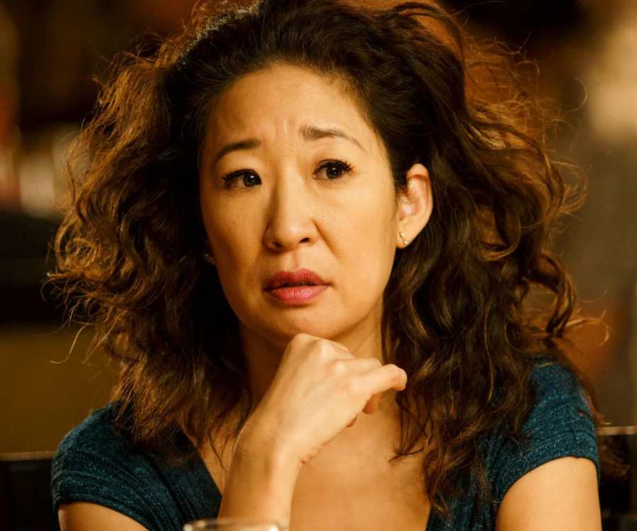 A woman with curly hair looks thoughtful, resting her chin on her hand, wearing a blue top.