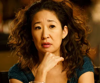 A woman with curly hair looks thoughtful, resting her chin on her hand, wearing a blue top.