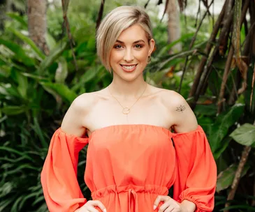 A woman in an orange off-shoulder dress with short hair stands in a lush green outdoor setting, smiling at the camera.