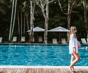 Pregnant woman in sunglasses walks by a pool with lounge chairs and umbrellas under tall trees.
