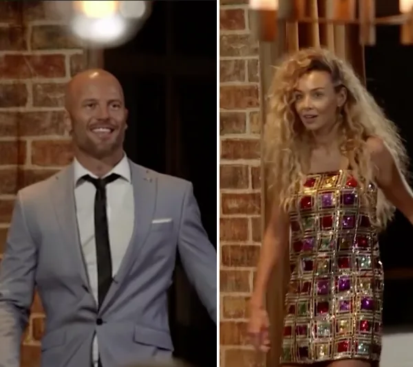 A man in a suit smiling beside a woman in a bejeweled dress with curly hair, both in a warmly lit room.