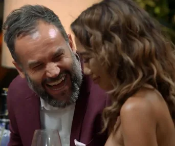 A man and woman share a light-hearted moment at dinner, both smiling and leaning toward each other.