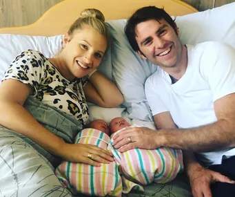Man and woman smiling in bed with two newborns wrapped in striped blankets.