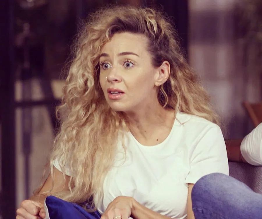 Woman with curly blonde hair looks surprised, wearing a white shirt, holding a blue pillow on a couch.