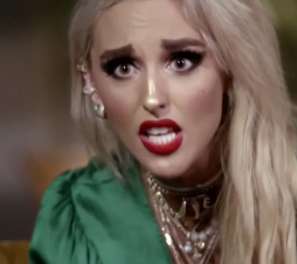 Blonde woman with dramatic makeup and red lips, wearing a green top, looks surprised or concerned, seemingly mid-conversation.