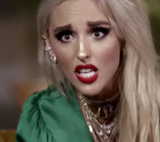 Blonde woman with dramatic makeup and red lips, wearing a green top, looks surprised or concerned, seemingly mid-conversation.