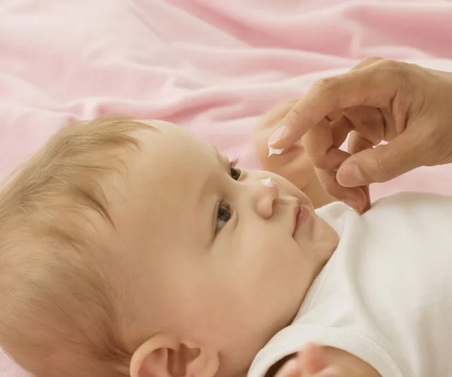 Baby lying down while an adult applies cream to its nose against a pink background.