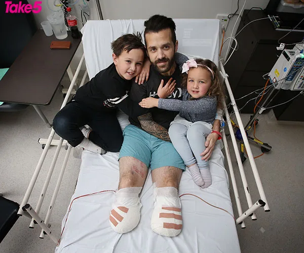 A man with bandaged feet sits in a hospital bed, embracing a young boy and girl, all smiling.
